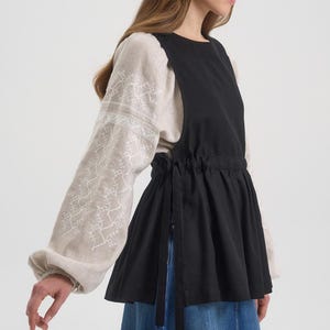 Op de afbeelding: Een zwarte top in schortstijl met een trekkoord in de taille en zijbanden, gelaagd over een beige blouse met geborduurde mouwen. De mouwen hebben een bloemenmotief. De outfit wordt gecompleteerd door een spijkerbroekrok.