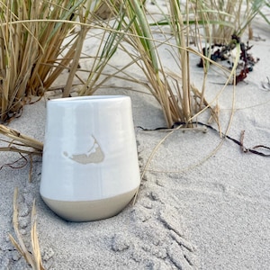 May include: A white ceramic tumbler with a beige base and a light beige outline of a landmass. The tumbler is sitting on a sandy beach with dune grass in the background. The tumbler is approximately 4 inches tall.