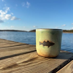 May include: A ceramic mug with a fish design. The mug is light green with a brown fish silhouette and a dark brown rim. It sits on a wooden dock with a lake and blue sky in the background.