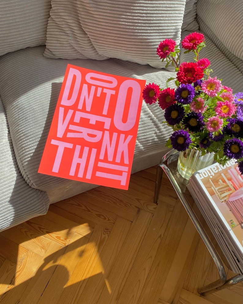 May include: A vibrant orange poster with the text "DON'T OVERTHINK THIS" in pink lettering. The poster is placed on a gray corduroy sofa. A bouquet of colorful flowers sits on a glass table, with magazines below.