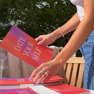 May include: A stack of books with a red and purple gradient cover. The text "DATE YOUR SELF" is printed in white. A person is holding one of the books. The background includes a wooden chair and green foliage.