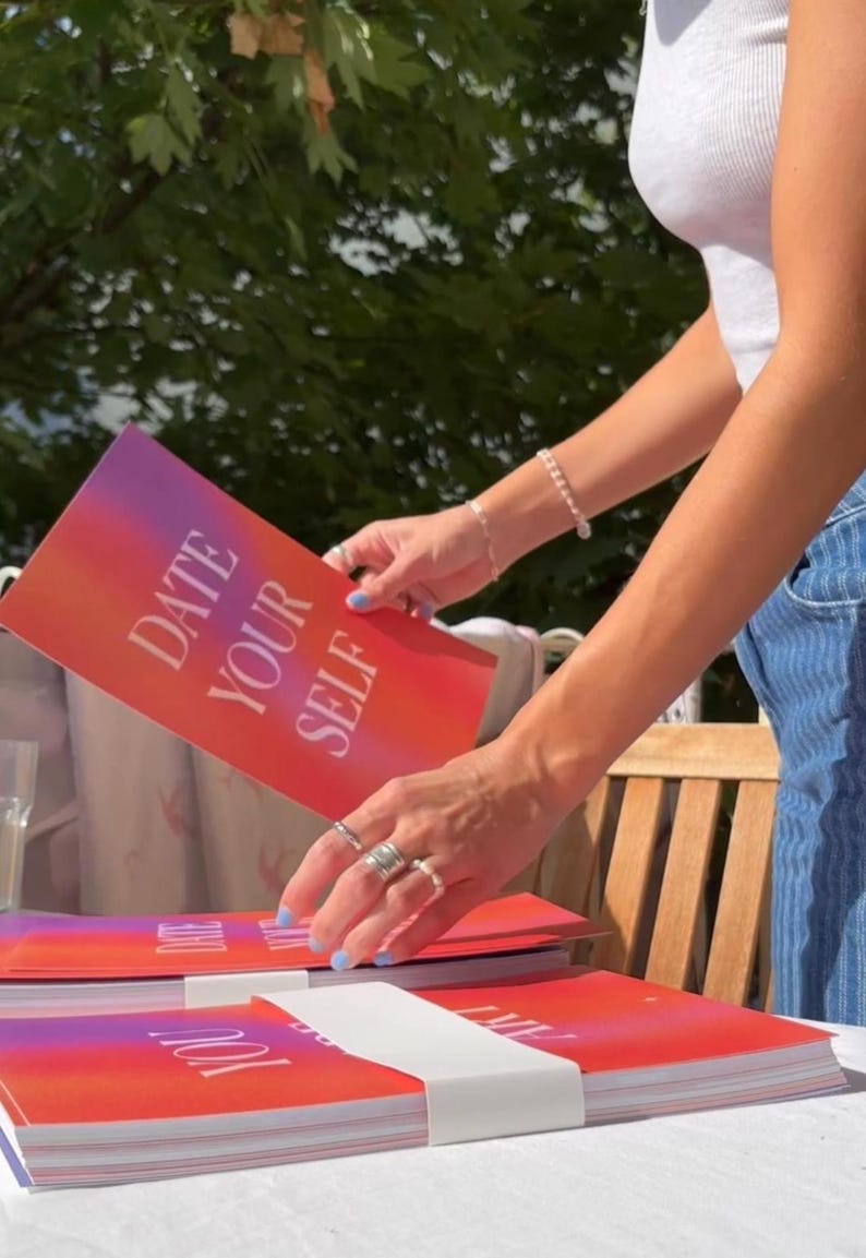 May include: A stack of books with a red and purple gradient cover. The text "DATE YOUR SELF" is printed in white. A person is holding one of the books. The background includes a wooden chair and green foliage.