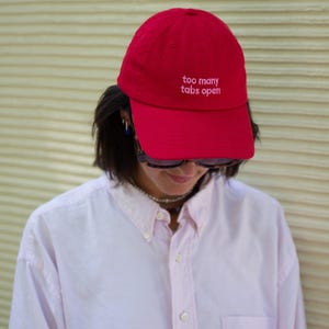 Könnte beinhalten: Rote Baseballkappe mit dem gestickten Text "too many tabs open". Getragen mit einem hellrosa Button-Down-Hemd.