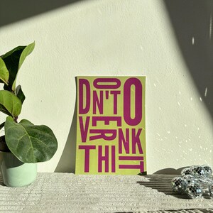 May include: A rectangular art print with the words "DON'T OVERTHINK THIS" in magenta block letters on a lime green background. A potted plant and a string of disco ball lights are in the background.
