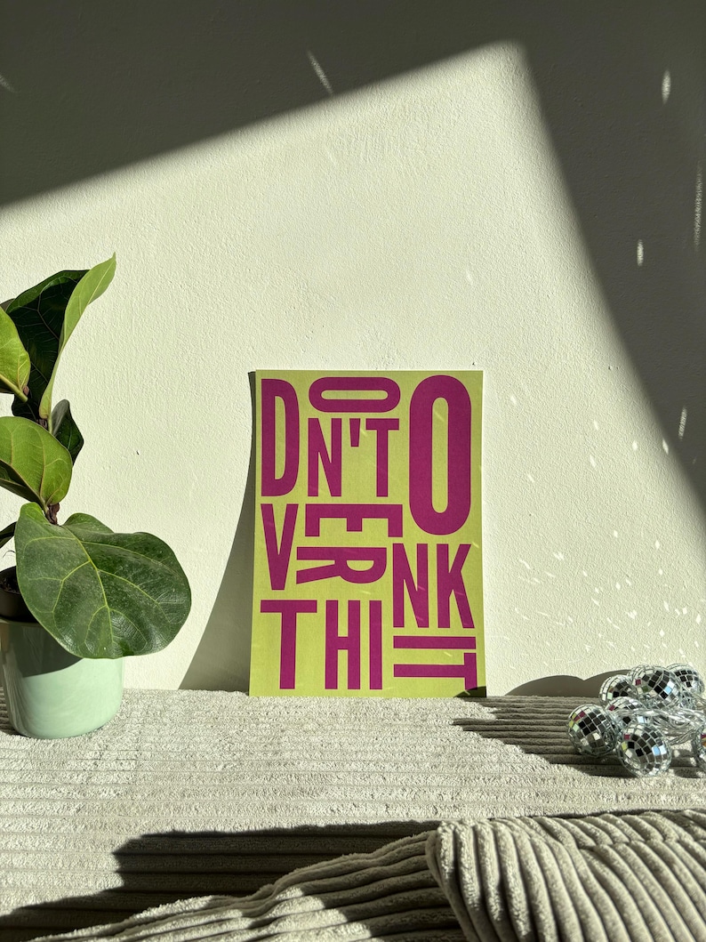 May include: A rectangular art print with the words "DON'T OVERTHINK THIS" in magenta block letters on a lime green background. A potted plant and a string of disco ball lights are in the background.