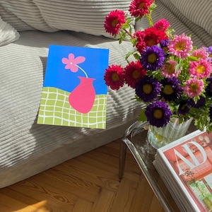 May include: A colorful still life featuring a vibrant bouquet of flowers in a glass vase, a magazine, and a framed artwork. The artwork depicts a pink vase with a flower against a blue background.