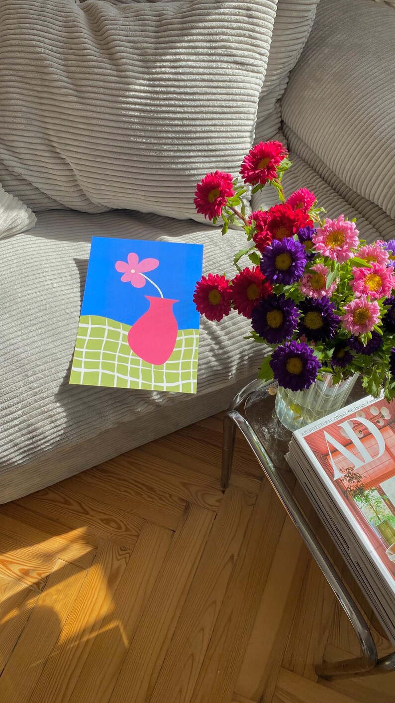May include: A colorful still life featuring a vibrant bouquet of flowers in a glass vase, a magazine, and a framed artwork. The artwork depicts a pink vase with a flower against a blue background.