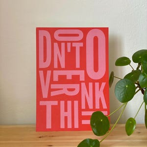 May include: A rectangular red art print with the words "DON'T OVERTHINK THIS" in pink block letters. The print is displayed on a wooden surface, with a green plant in the background.