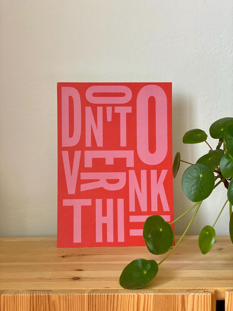 May include: A rectangular red art print with the words "DON'T OVERTHINK THIS" in pink block letters. The print is displayed on a wooden surface, with a green plant in the background.
