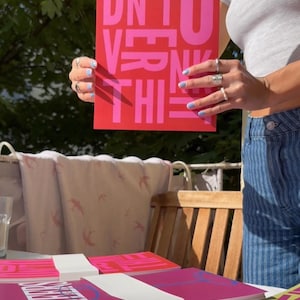 May include: A red poster with the words "DON'T OVERTHINK THIS" in pink block letters. Several stacks of similar posters are on a table. The image is taken outdoors in natural light.