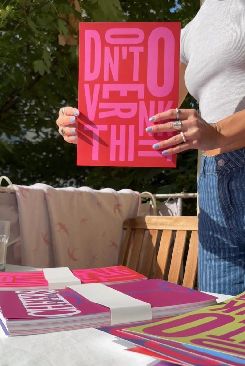 May include: A red poster with the words "DON'T OVERTHINK THIS" in pink block letters. Several stacks of similar posters are on a table. The image is taken outdoors in natural light.
