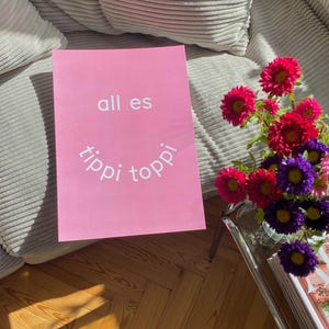 May include: A pink poster with the words "all es tippi toppi" in white lettering. A vase of colorful flowers sits on a glass table next to the poster. The flowers are red, purple, and pink.
