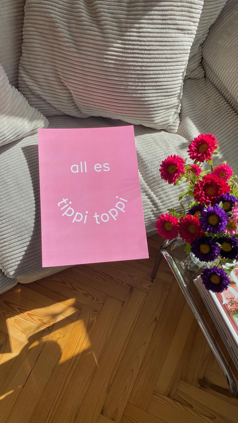 May include: A pink poster with the words "all es tippi toppi" in white lettering. A vase of colorful flowers sits on a glass table next to the poster. The flowers are red, purple, and pink.