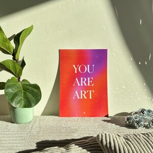 May include: A colorful art print with the words "YOU ARE ART" in white lettering. The background transitions from red to purple. A potted plant and decorative disco ball are also in the image.