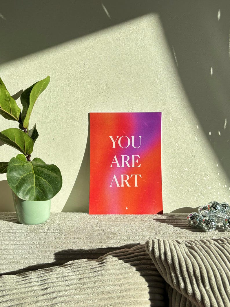 May include: A colorful art print with the words "YOU ARE ART" in white lettering. The background transitions from red to purple. A potted plant and decorative disco ball are also in the image.