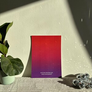May include: A rectangular card with a gradient of red to purple, featuring the text "sex is nice but have you tried conversation?" The card is displayed on a textured surface, with a plant and disco ball lights in the background.