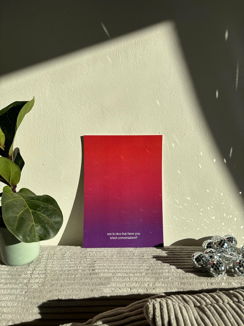 May include: A rectangular card with a gradient of red to purple, featuring the text "sex is nice but have you tried conversation?" The card is displayed on a textured surface, with a plant and disco ball lights in the background.