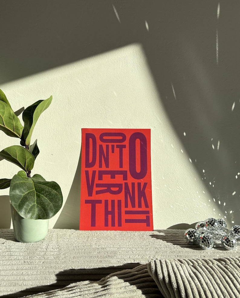 May include: A red rectangular art print with the words "DON'T OVERTHINK IT" in purple block letters. A small potted plant with large green leaves and a decorative disco ball are also visible.