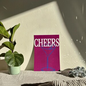 May include: A magenta print with the word "CHEERS" in white and a blue cocktail glass illustration. The print is displayed on a corduroy surface, with a small potted plant and disco ball accents.