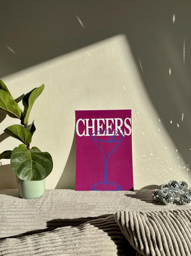 May include: A magenta print with the word "CHEERS" in white and a blue cocktail glass illustration. The print is displayed on a corduroy surface, with a small potted plant and disco ball accents.