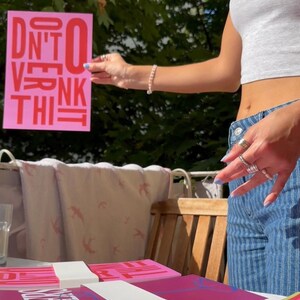 May include: A stack of pink and magenta notebooks with white bands, and a pink sign that says "DON'T OVERTHINK IT" in red. The image is taken outdoors on a sunny day.