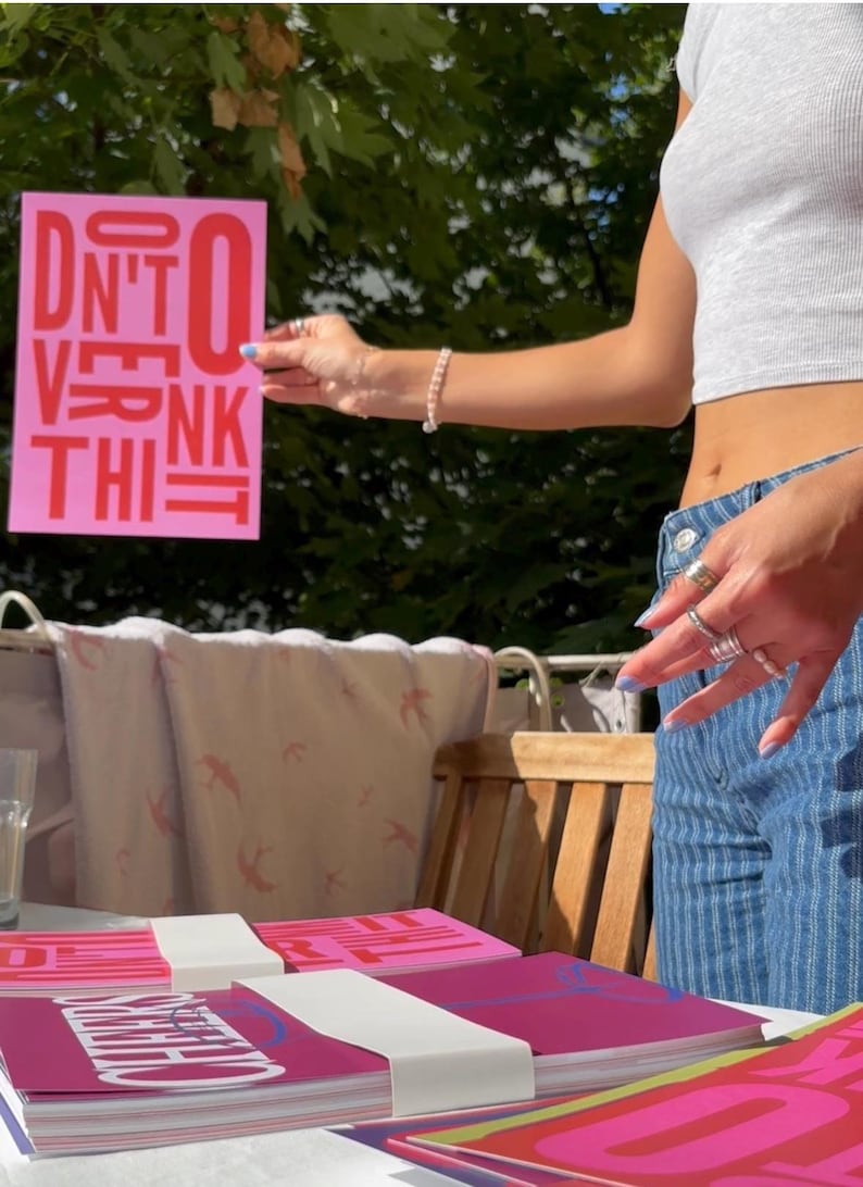 May include: A stack of pink and magenta notebooks with white bands, and a pink sign that says "DON'T OVERTHINK IT" in red. The image is taken outdoors on a sunny day.