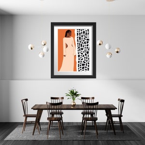 May include: A black framed print of a woman in a white dress standing in front of a black and white geometric pattern. The print is hanging on a white wall above a wooden dining table with four chairs.