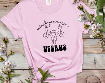Peut inclure: T-shirt rose avec un graphique noir d'un utérus avec les doigts d'honneur levés et le texte "mind your own uterus" dans une police rétro.