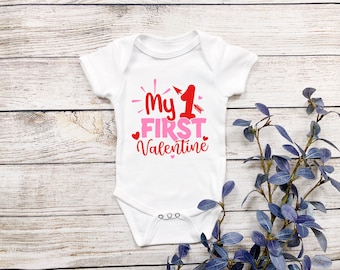 May include: White baby onesie with the text "My 1st Valentine" in red and pink lettering with a red arrow and hearts.