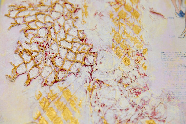 May include: Close-up of a mixed media art piece with gold and white paint and a lace overlay. The background is a light pink with a faint text pattern.