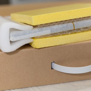 May include: A brown cardboard box with a white handle. The box is filled with yellow foam padding and a white protective layer.