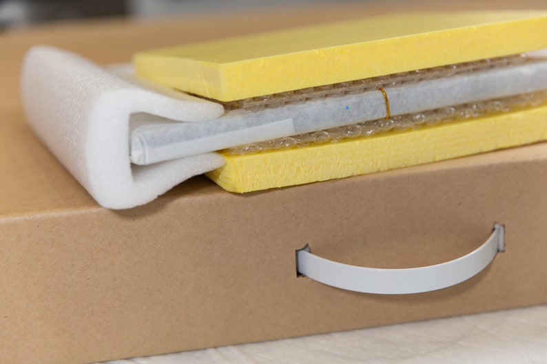 May include: A brown cardboard box with a white handle. The box is filled with yellow foam padding and a white protective layer.