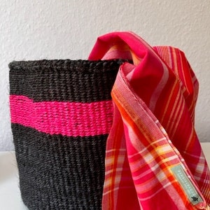 Könnte beinhalten: Ein gewebter schwarzer Korb mit einem horizontalen Band in leuchtendem Pink. Ein rot-rosa karierter Schal mit Fransen ist über die Seite des Korbs drapiert. Der Schal hat ein kleines Etikett.