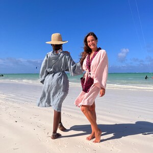 May include: Two people on a sandy beach wearing robes. One person wears a light blue robe with a straw hat. The other person wears a pink robe with a patterned collar. The sky is blue, and the ocean is visible in the background.