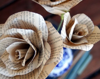 Puede incluir: Tres rosas de papel hechas con las páginas de libros antiguos. Las rosas están dispuestas en un ramo y se encuentran en un jarrón azul y blanco. Las rosas son de color marrón claro y tienen un aspecto vintage.
