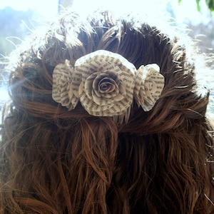 Peut inclure: Trois fleurs en papier faites de pages de livres sont disposées en rangée et épinglées sur les cheveux bruns d'une femme. Les fleurs sont blanches avec du texte noir.