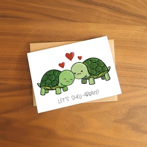 May include: A white greeting card with two cartoon turtles, green shells, and smiling faces. Red hearts float above them. The text "LET'S SHELL-ABRATE!" is printed below the turtles. The card rests on a brown envelope.