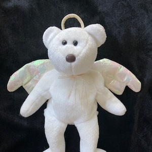 Può includere: Orso di peluche bianco a forma di angelo con ali iridescenti e un'aureola. L'orso ha un naso marrone e occhi neri.