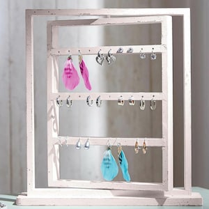 Può includere: Un porta orecchini in legno bianco con tre livelli di ganci per appendere gli orecchini. Il supporto ha una finitura bianca invecchiata e presenta una varietà di orecchini, tra cui orecchini a piuma, orecchini a cerchio e orecchini a perno.