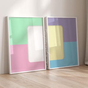 May include: Two framed abstract art prints. One features a green, pink, and white color block design. The other print has a yellow, purple, and blue color block design. Both are in white frames and lean against a white wall.