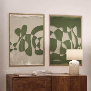 Puede incluir: Dos impresiones artísticas abstractas enmarcadas con diseños geométricos verdes y beige cuelgan sobre un mueble de madera oscura. Una lámpara blanca con diseño de rostro está sobre el mueble. Una mesa blanca y velas están en primer plano.