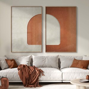May include: Two rectangular wall art prints in thin wooden frames, featuring a split composition of off-white and terracotta orange colors. The design incorporates a large arch shape and vertical line textures on the orange sections.
