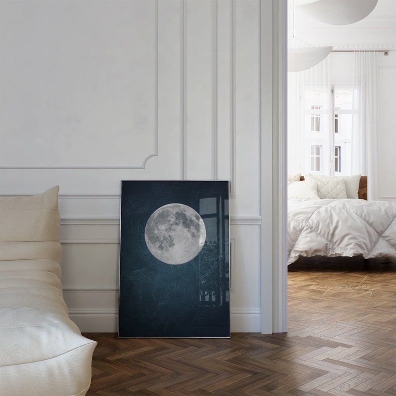 May include: A framed print of a full moon in a dark blue sky. The moon is white and has a textured surface. The print is hanging on a white wall in a room with a white couch and a doorway leading to another room.
