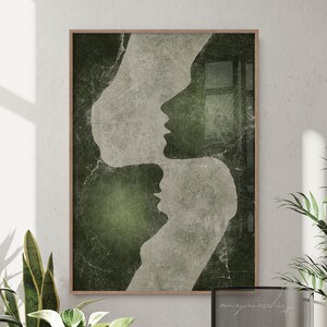 May include: Abstract art print showing three overlapping profiles in white against a textured, dark green and grey background. The artwork is framed in a natural wood tone, creating a sense of depth. A decorative piece of wall art.