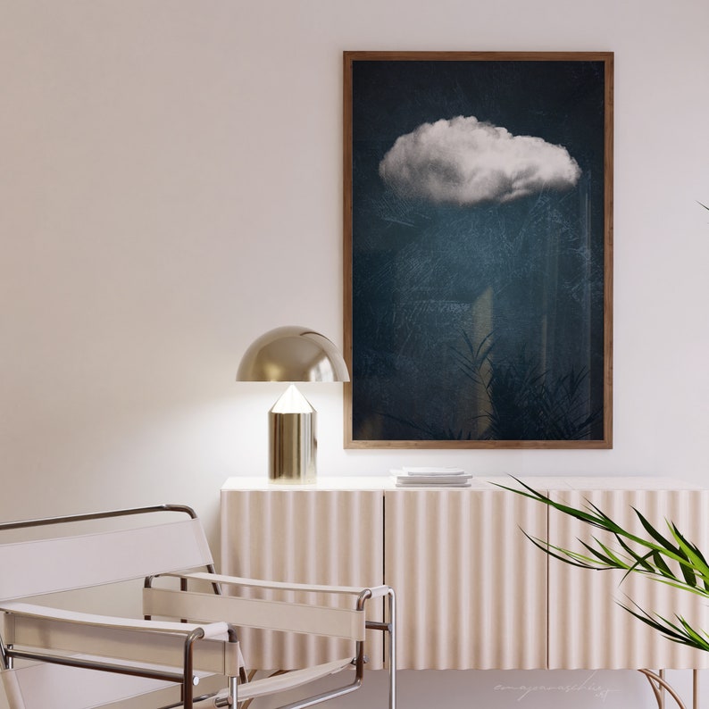 May include: A white cloud against a dark blue background in a framed print hanging on a white wall above a light beige cabinet with a gold lamp and a white chair.