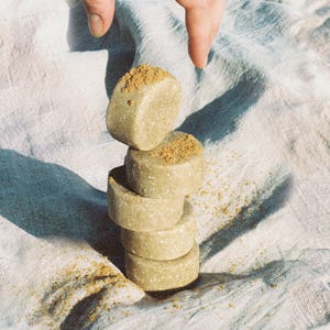May include: A stack of four round, light brown soap bars with a light brown powder sprinkled on top. The soap bars are stacked on a white fabric surface.