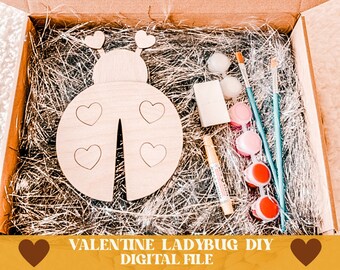 Puede incluir: Un kit de manualidades de madera en forma de mariquita con pintura, pinceles y pegamento. La mariquita tiene una forma de corazón en sus alas. El kit viene en una caja marrón con una tarjeta blanca que dice "Feliz Día de San Valentín".
