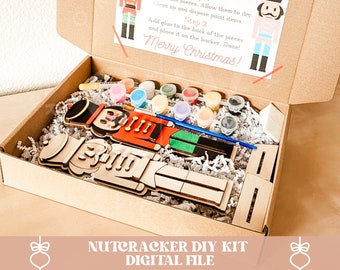 May include: A brown cardboard box with a Nutcracker DIY kit inside. The kit includes a wooden nutcracker, paint, brushes, and a backing board. The box has a printed label with instructions and the text "Nutcracker DIY Kit" and "Merry Christmas!"