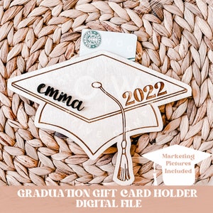 May include: A wooden graduation cap gift card holder with the name "emma" and the year "2022" engraved on it. The cap is sitting on a woven surface.
