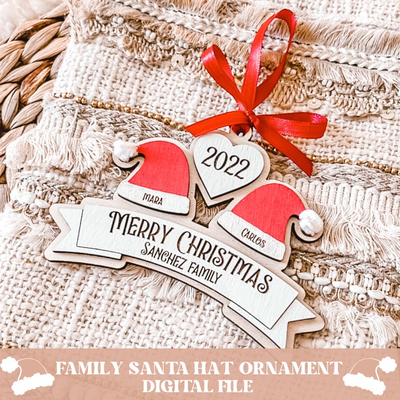 Puede incluir: Un adorno navide&ntilde;o de madera con dos gorros de Pap&aacute; Noel rojos con pompones blancos, un coraz&oacute;n con el a&ntilde;o 2022 y una pancarta que dice "Feliz Navidad Familia Sanchez".
